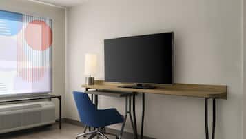 A modern hotel room with a flat-screen TV on a wooden desk, a blue office chair, and a large window with abstract art.