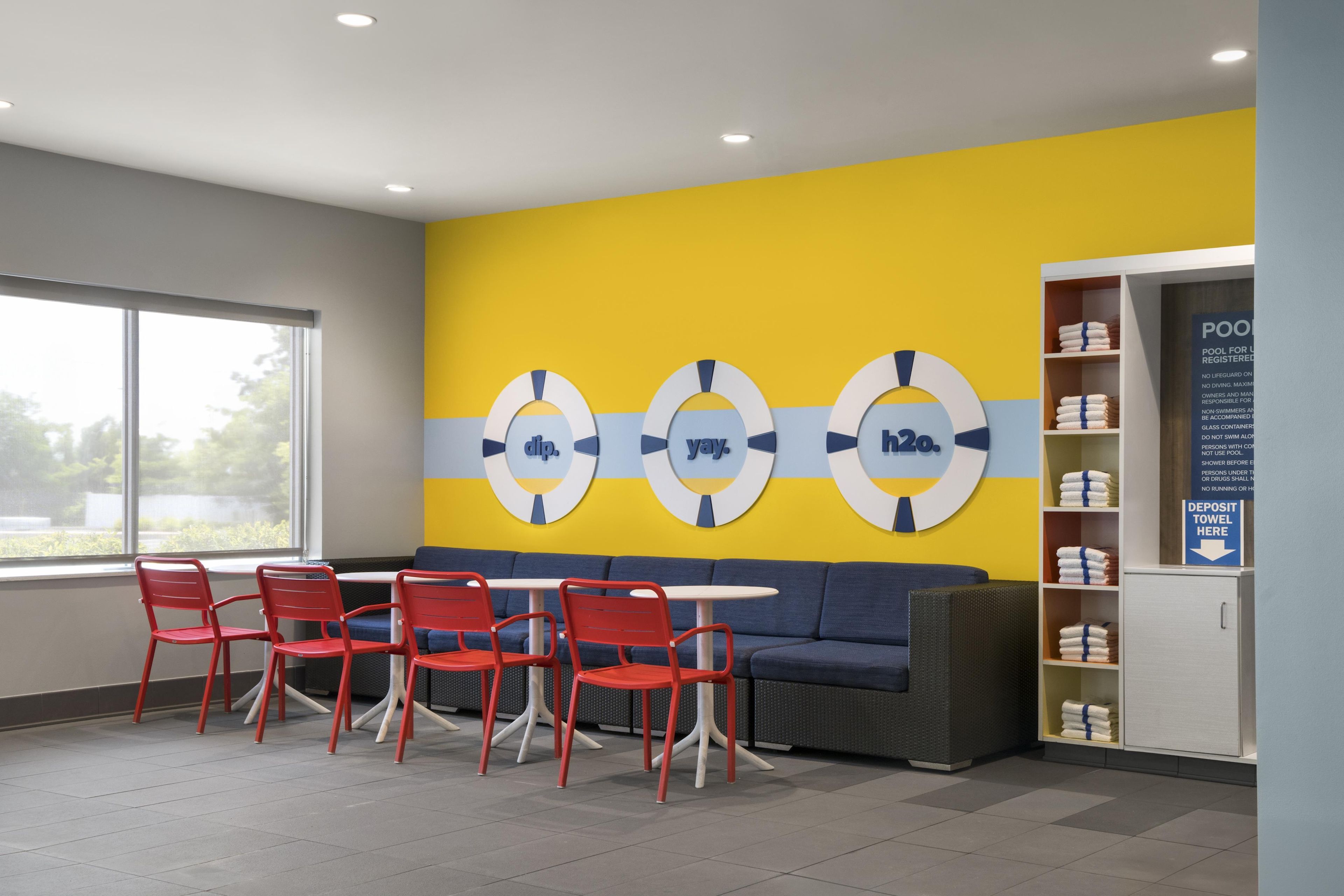 Un espacio interior moderno con una paleta de colores amarillo y gris, que incluye una zona de estar con sillas rojas, un sofá gris y una piscina con un cartel que indica que es solo para usuarios registrados.
