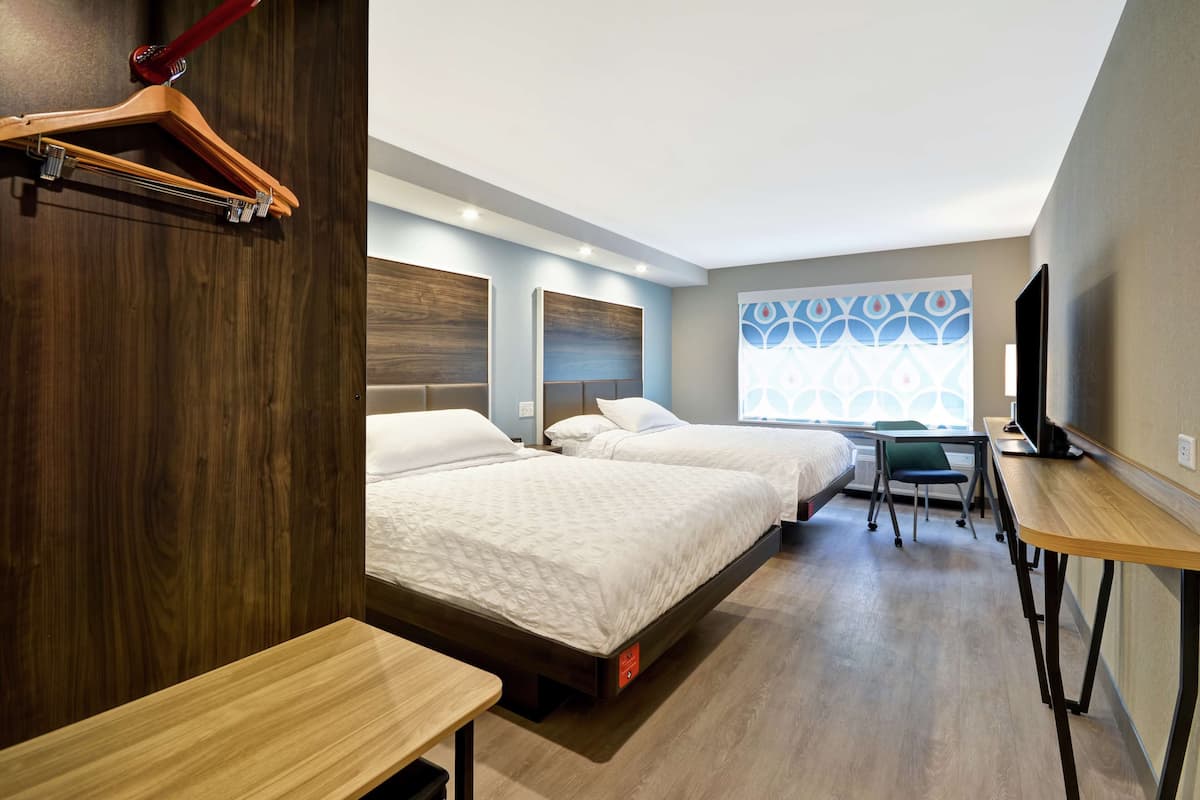 Habitación de hotel con dos camas, un escritorio, una silla, un televisor y un ventanal con un diseño geométrico.