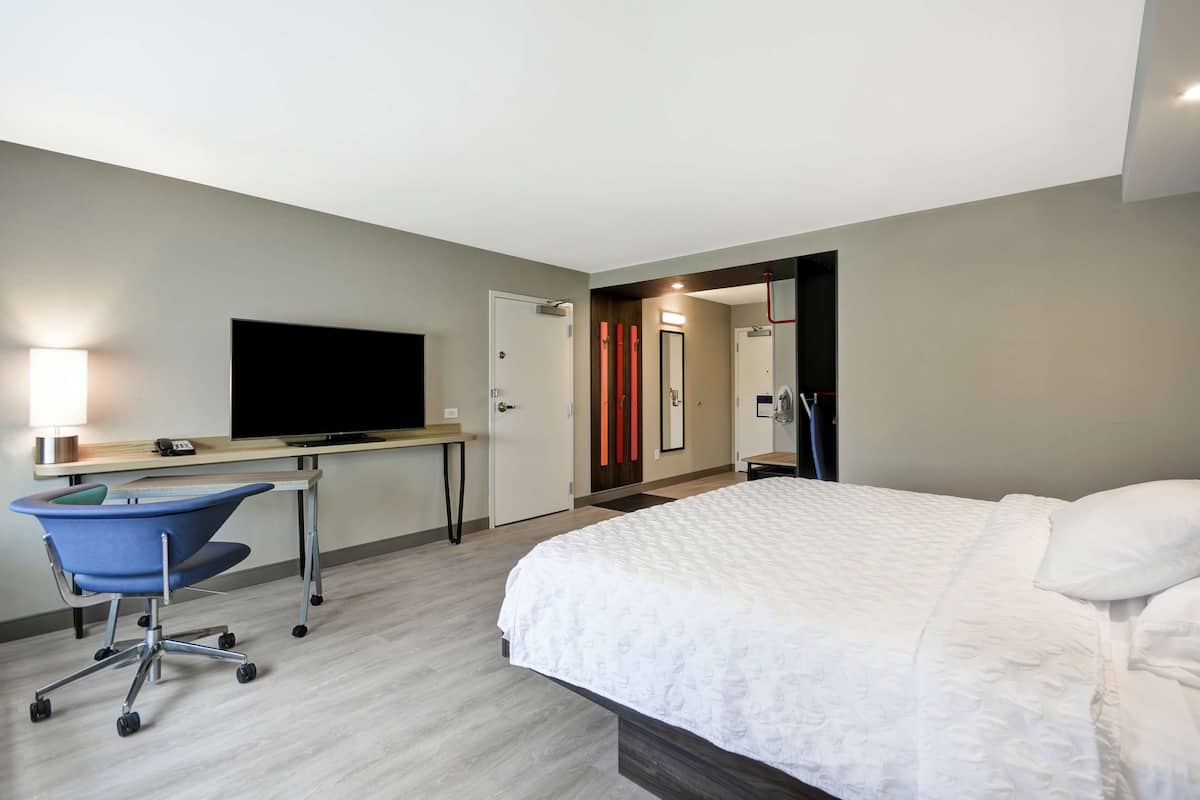 Habitación de hotel con cama, escritorio, silla, televisor y espejo.