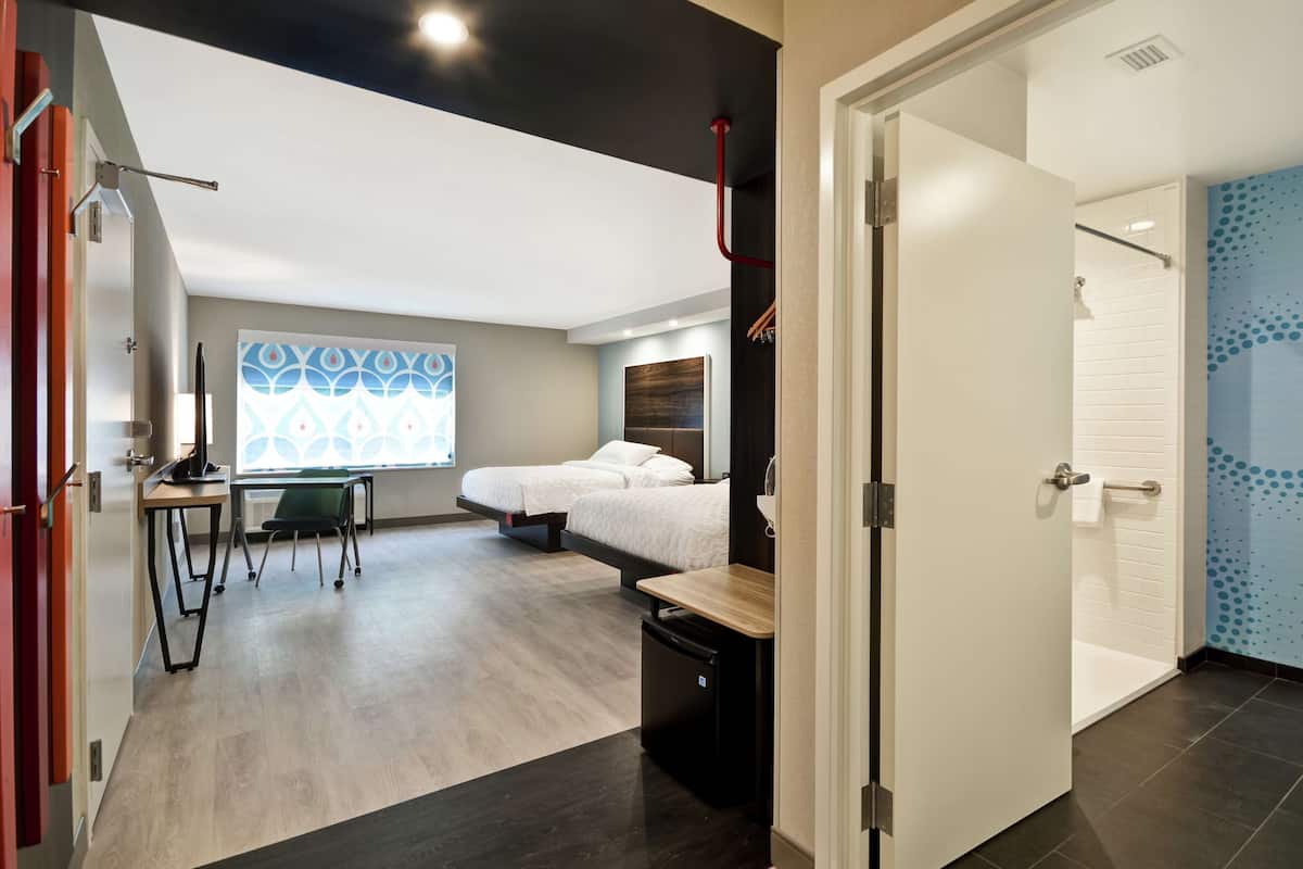 Una habitación de hotel moderna con cama, escritorio, silla, una kitchenette pequeña y un baño con ducha e inodoro.