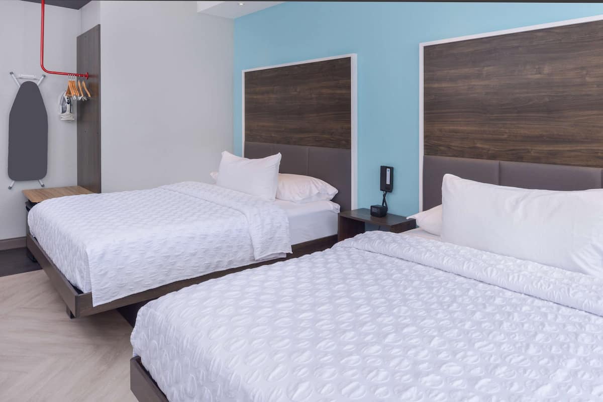 A hotel room with two beds, a wooden headboard, a small table, and a wall-mounted ironing board.