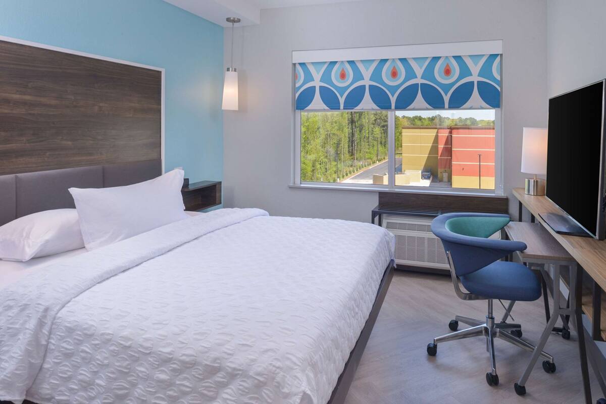 A hotel room with a bed, a desk with a chair, a TV, and a window with a view of trees and buildings.