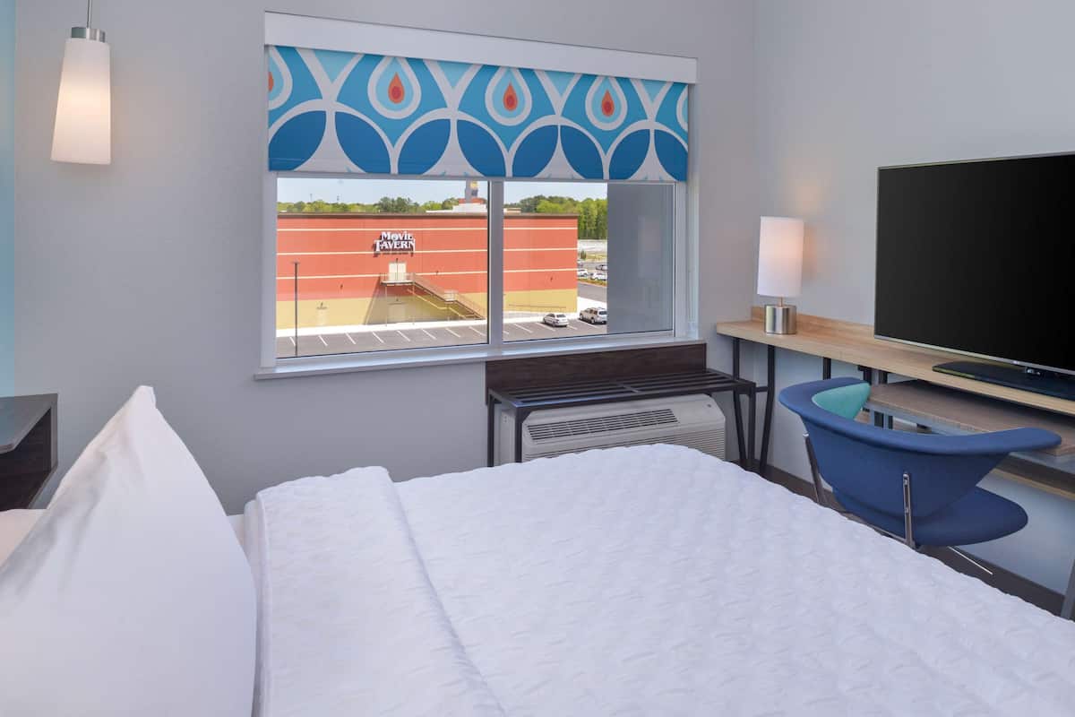A hotel room with a bed, a desk with a computer, a chair, a TV, and a window with a view of a building outside.