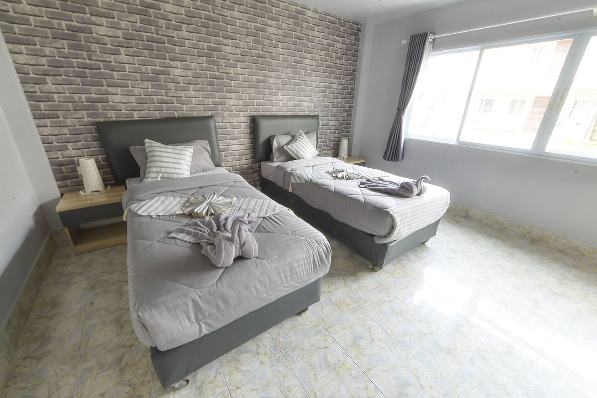 A bedroom with two beds, a brick wall, a window, and a nightstand with a lamp.
