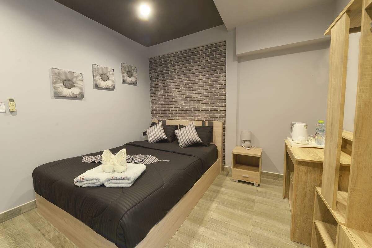 A modern bedroom with a large bed, a wooden headboard, a brick-patterned accent wall, and a small bedside table with a kettle.