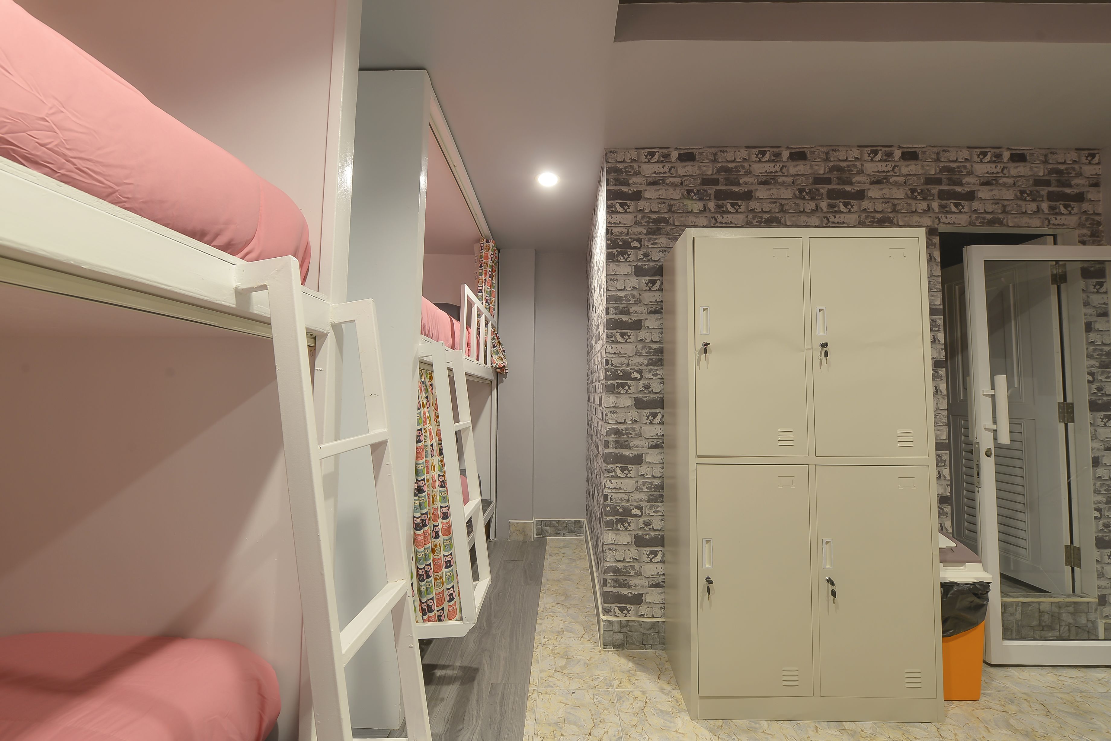 Un lit superposé avec une literie rose, une armoire avec un mur décoré d’un motif imitant des briques, et une porte donnant sur une autre pièce.