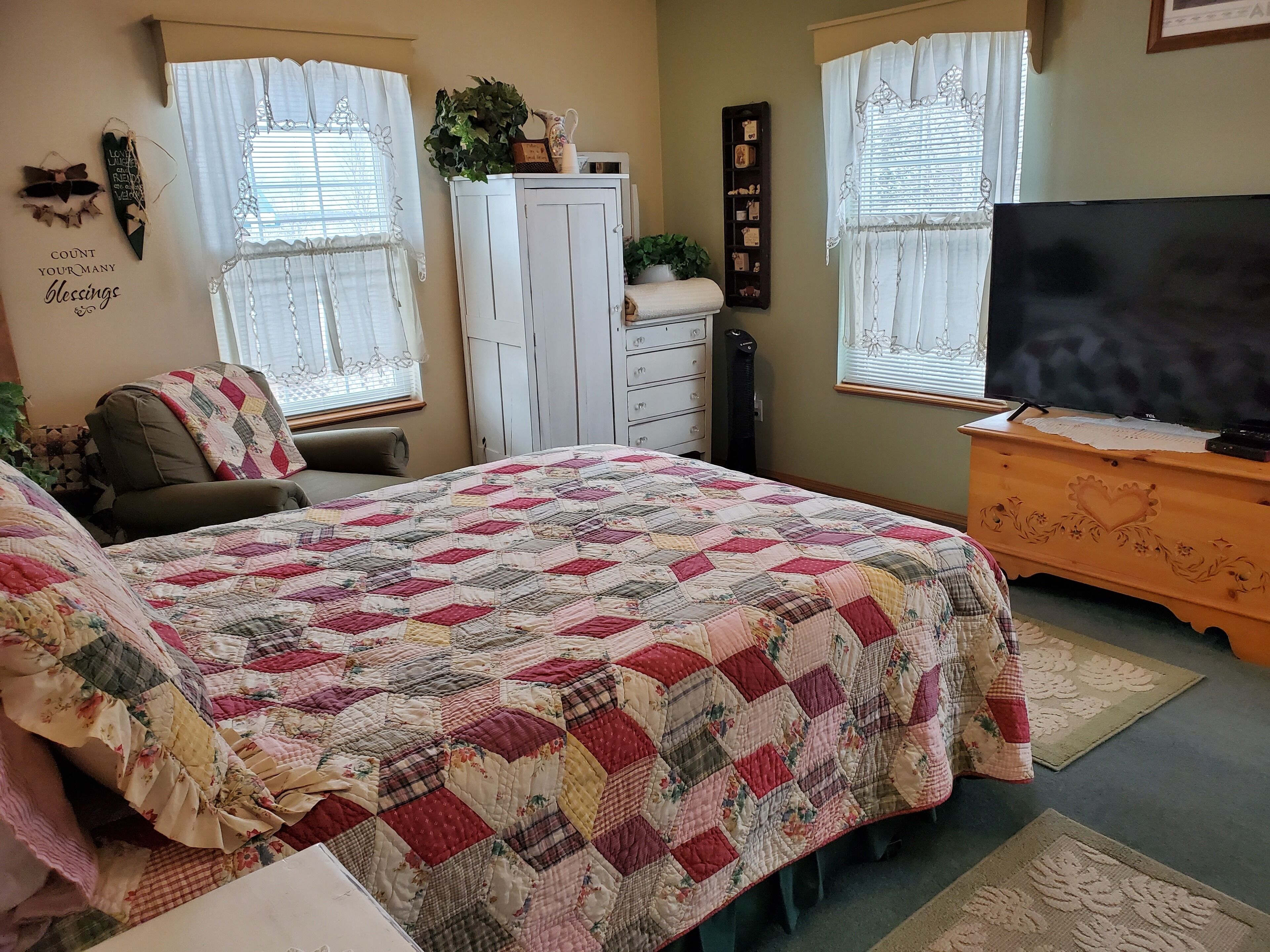 A bedroom with a large bed, a chair, a dresser, a television, and two windows with curtains.