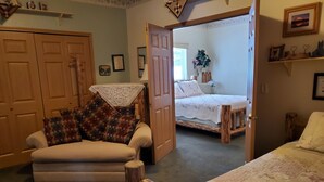 A cozy room with a sofa, a wooden bench, and a bed with a lace cover. There are framed pictures on the walls and a plant on the bed. - Alaska's Lake Lucille B&B (Wasilla)