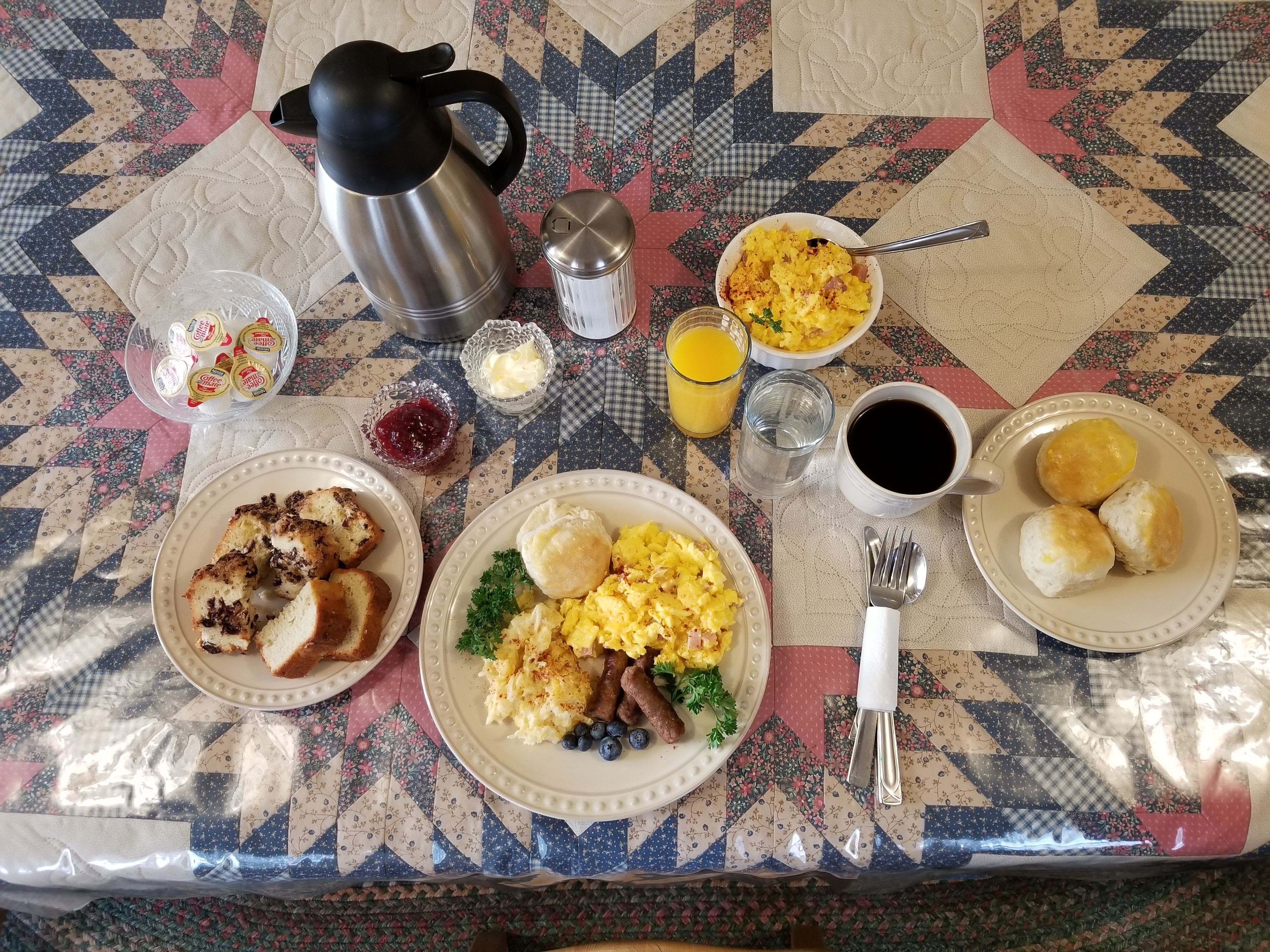A breakfast table with a coffee pot, butter, jam, juice, coffee, and biscuits.