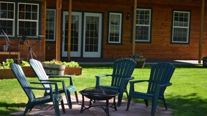 A patio with four Adirondack chairs and a round table, surrounded by a wooden house with large windows and a brick pathway. - Alaska's Lake Lucille B&B (Wasilla)