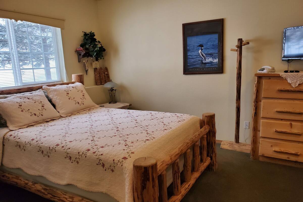 A bedroom with a wooden bed frame, a bedside table with a lamp, a dresser, a framed picture on the wall, and a window with blinds.