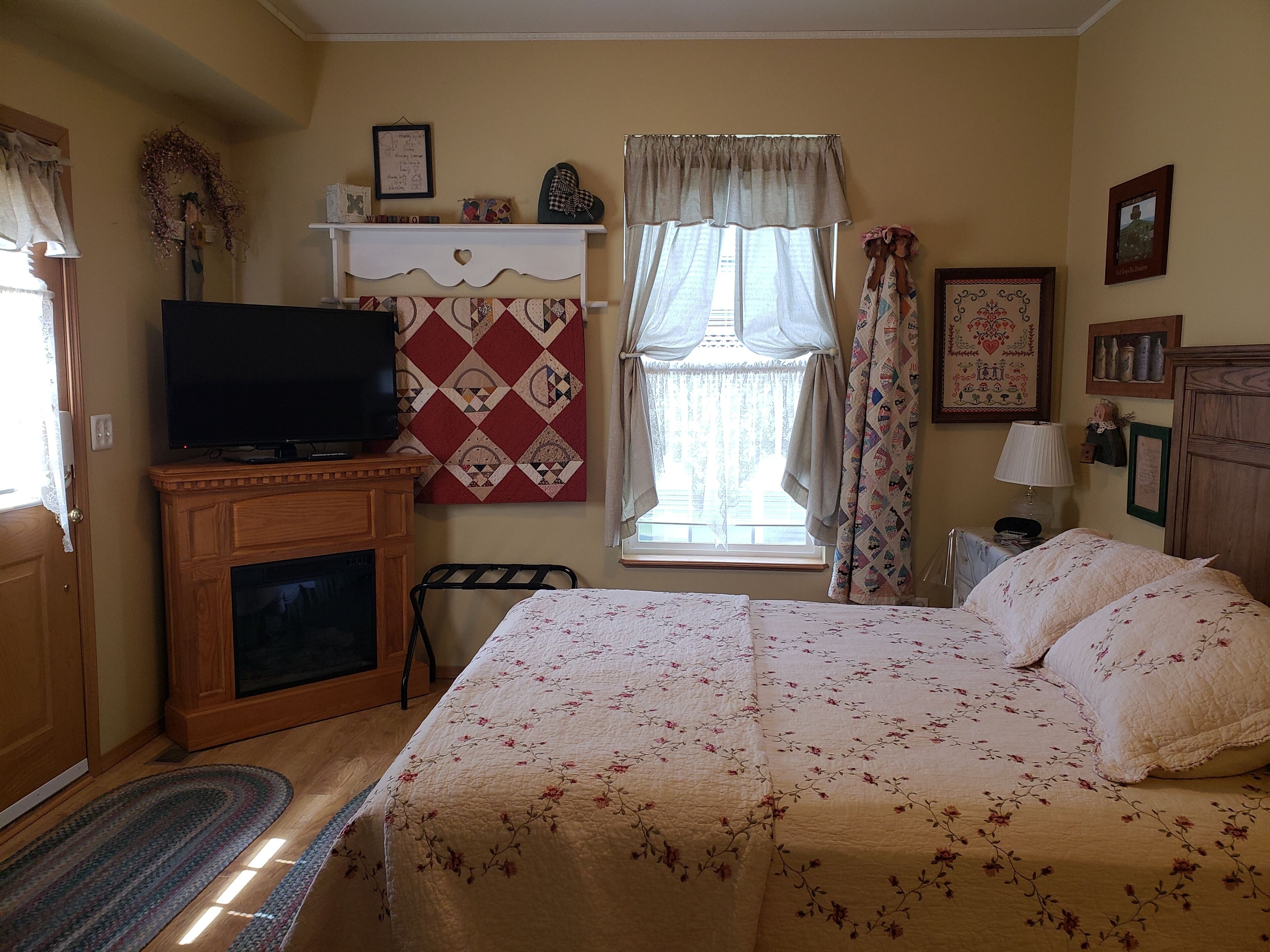 A bedroom with a bed, a television, a fireplace, a window with curtains, and a wall decorated with pictures and a quilt.