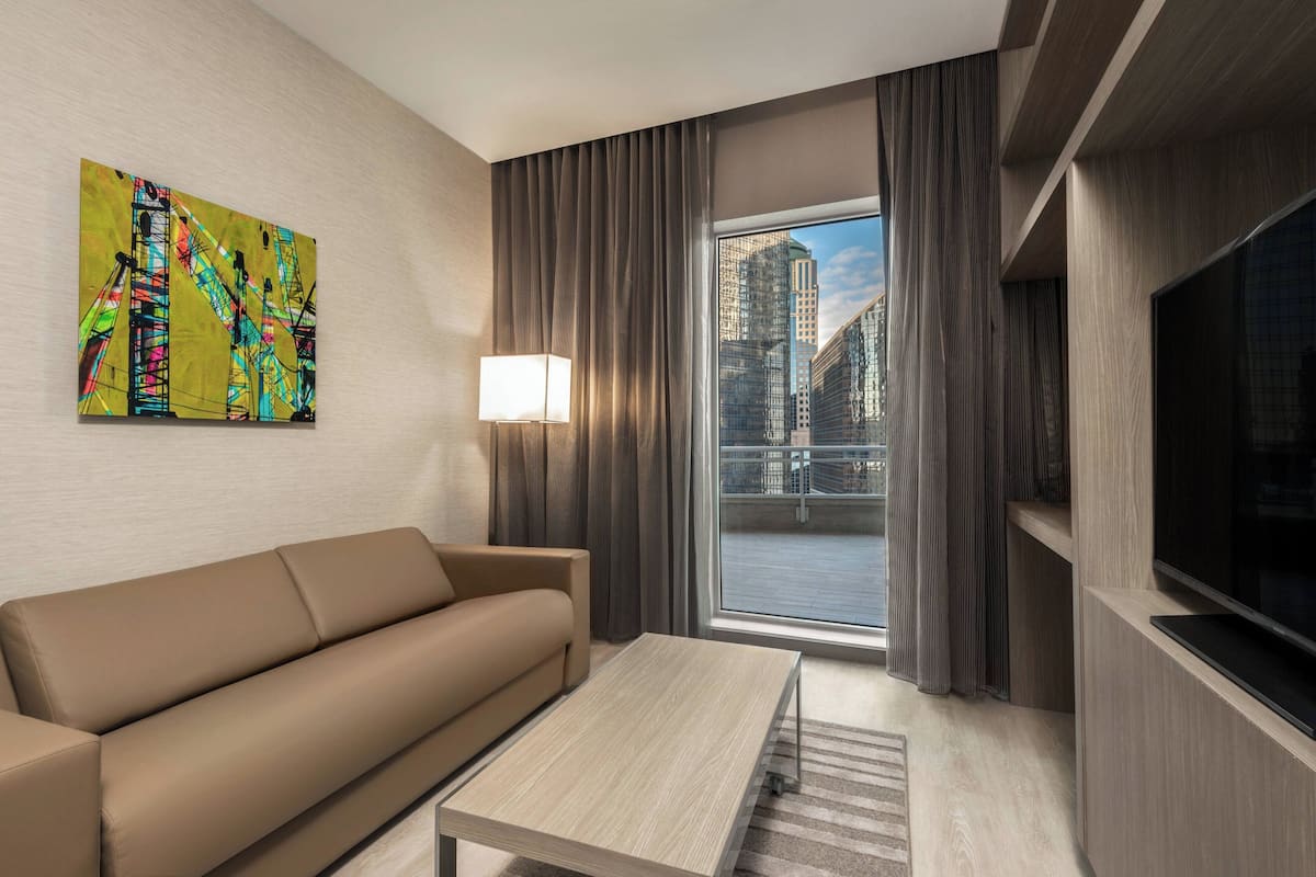 A modern hotel room with a sofa, a coffee table, a TV, a large window with curtains, and a cityscape view.