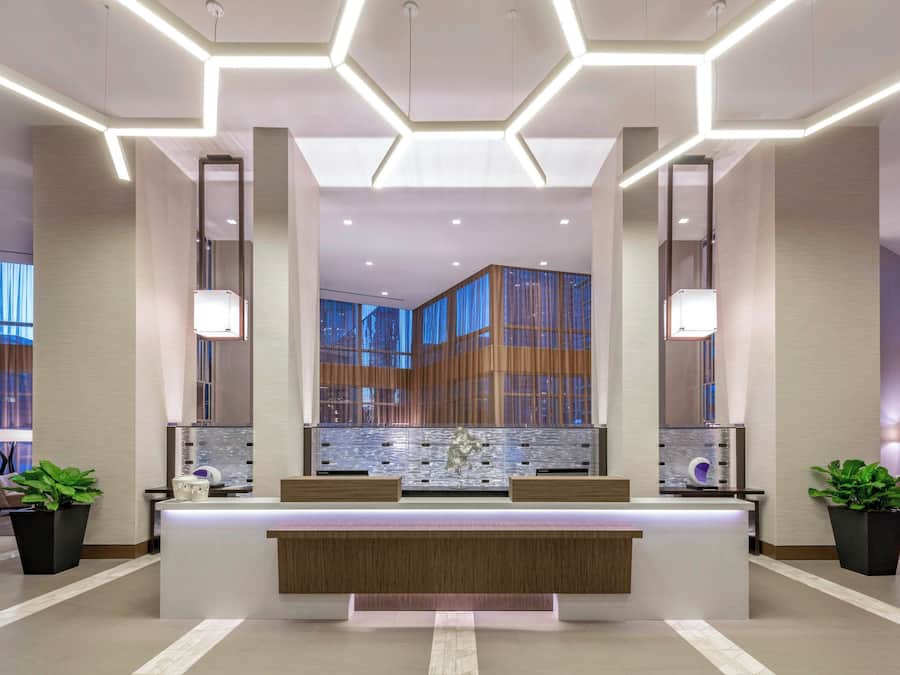 Een moderne hotellobby met een receptiebalie, grote ramen en een geometrisch plafondontwerp.