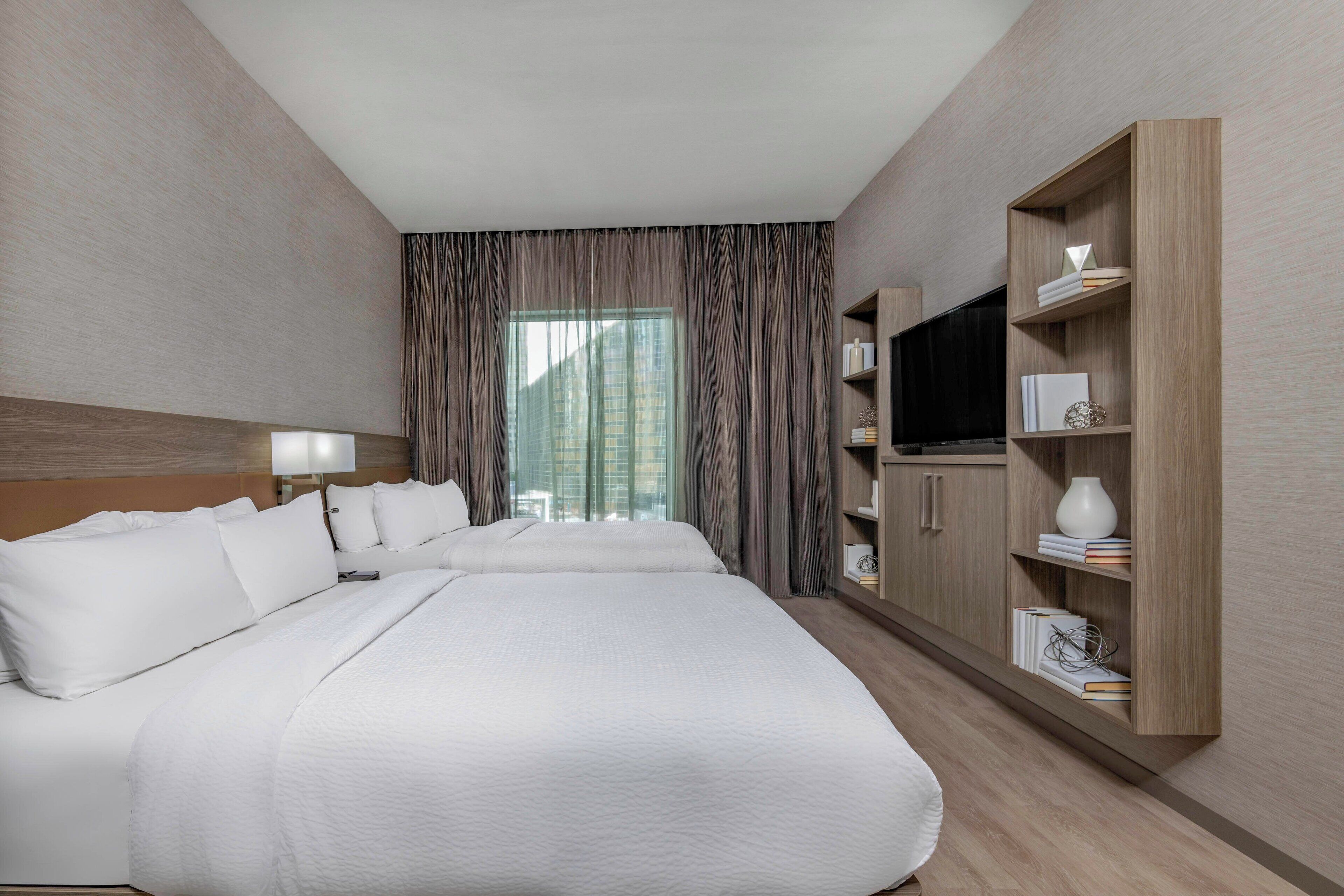 A hotel room with a large bed, a TV, a bookshelf, and a window with curtains.