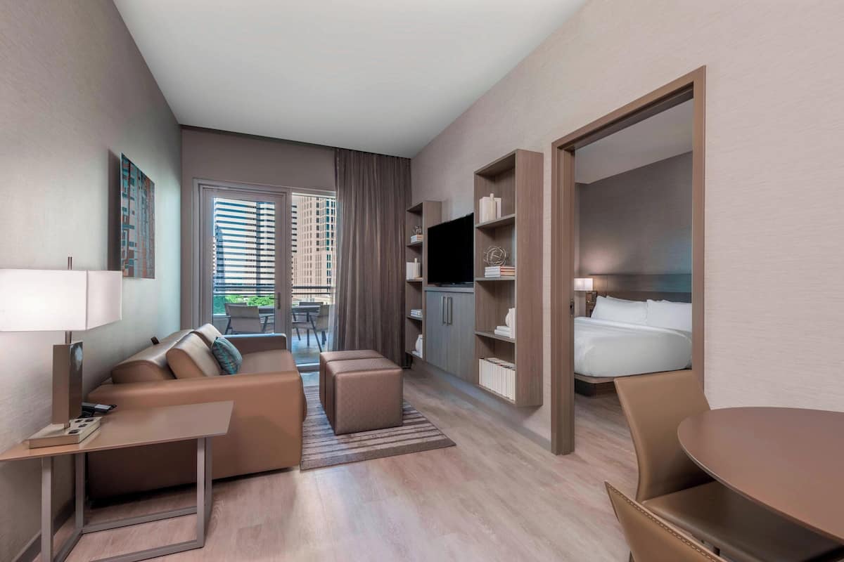 A modern hotel room with a sofa, ottoman, TV, and a dining area.