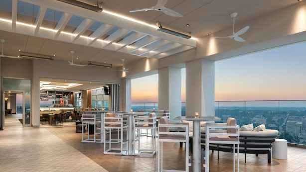 A modern bar area with high tables and chairs, a seating area with sofas, and large windows offering a city view. - AC Hotels by Marriott Charlotte City Center (Charlotte)