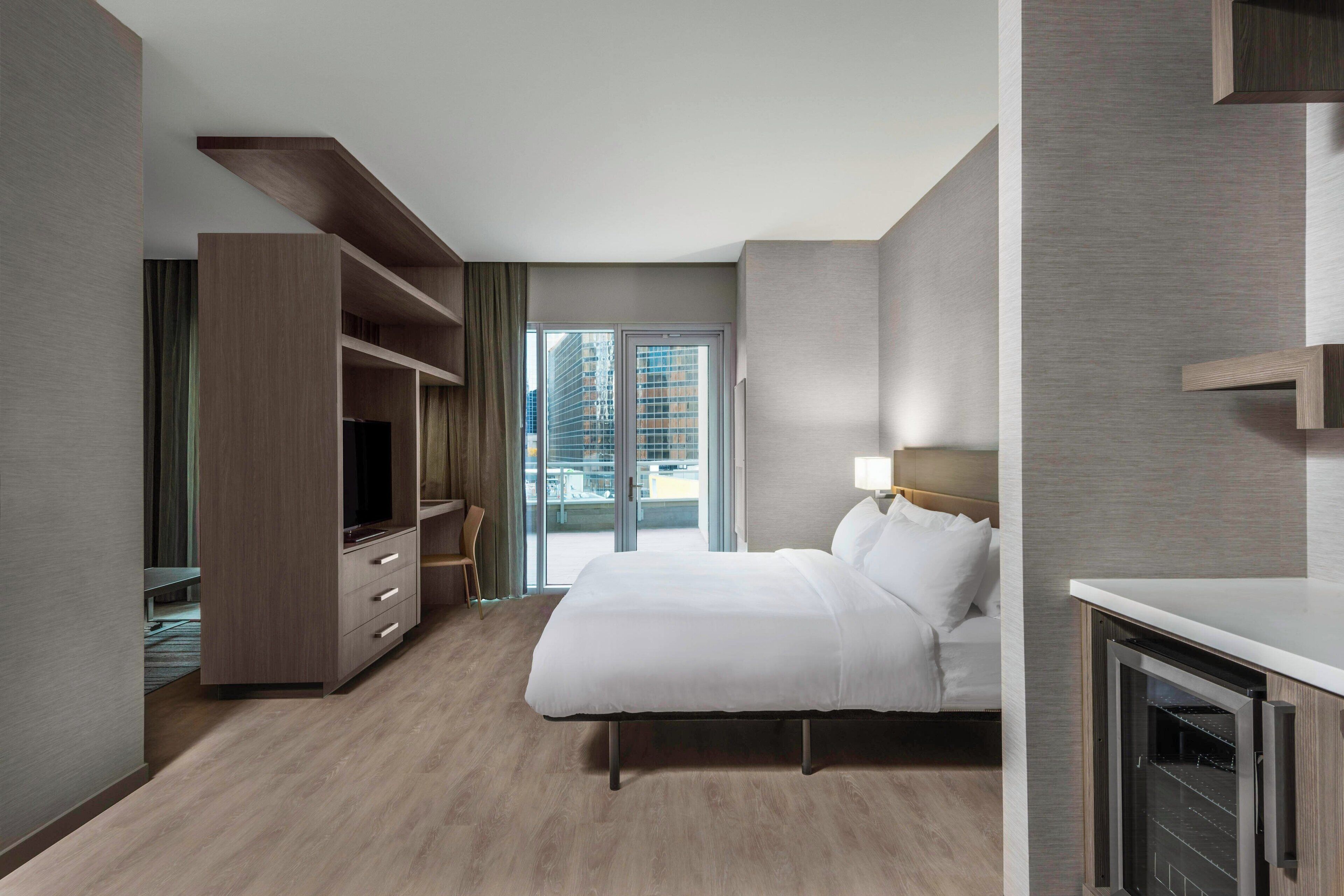 A modern hotel room with a large bed, a TV, a desk, and a view of the city through the window.