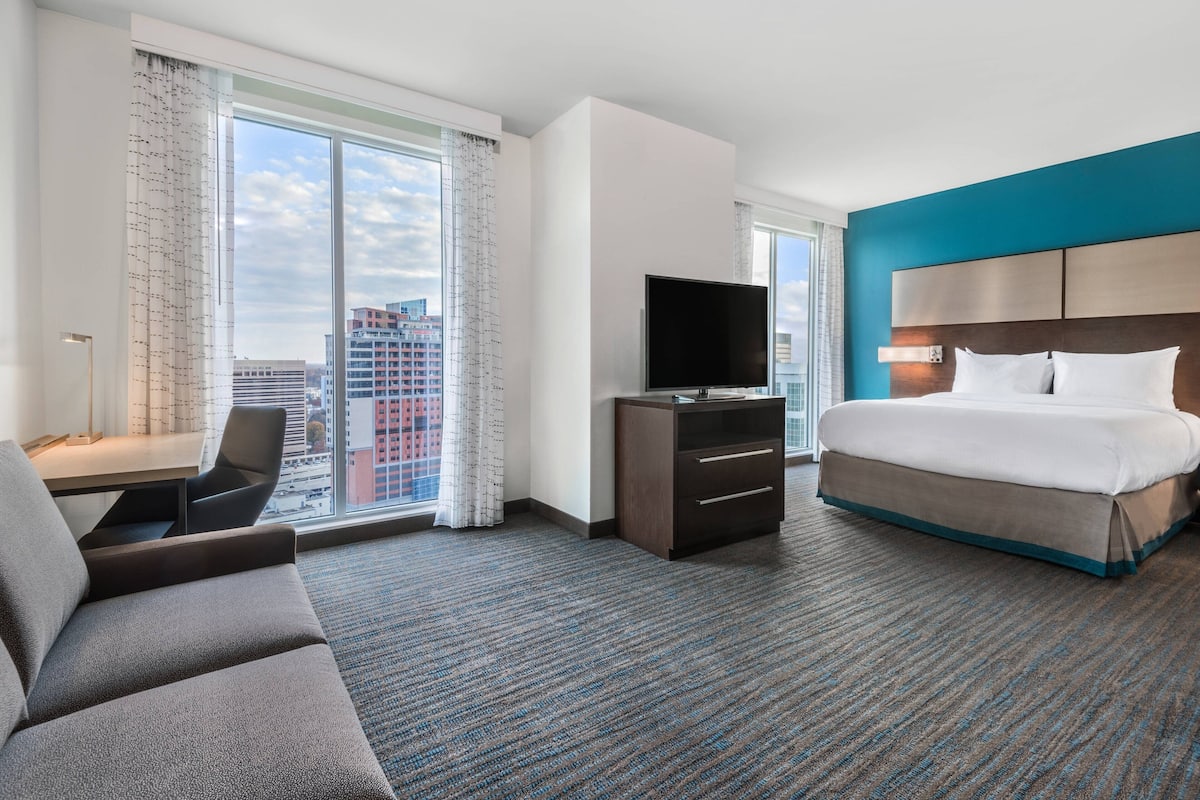 A hotel room with a large bed, a desk with a chair, a television, and a view of the city through large windows.