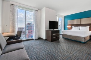 Habitación de hotel con una cama grande, un escritorio con silla, televisor y vistas a la ciudad a través de amplias ventanas.