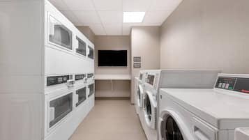 A laundry room with multiple washing machines and dryers, a flat-screen TV mounted on the wall, and a sign indicating cycle times.