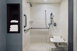 A modern bathroom with a walk-in shower, grab bars, a bench, and a shelf with neatly folded towels.