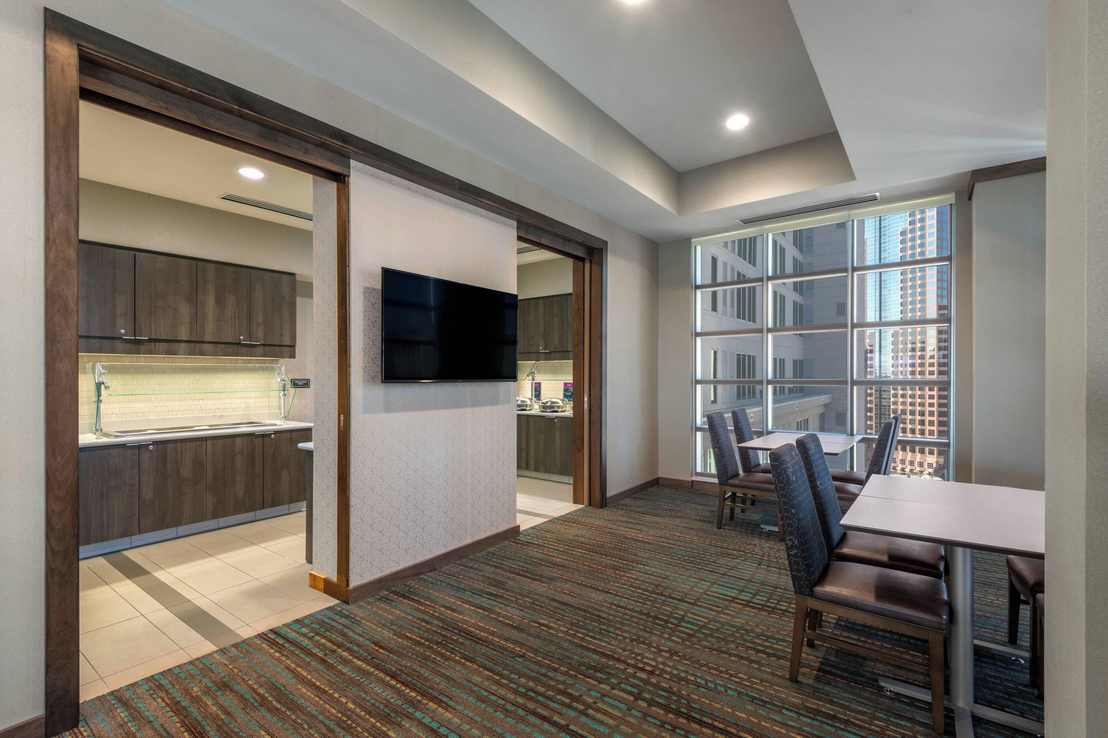 Habitación de hotel con kitchenette, televisor de pantalla plana, zona de comedor con mesa y sillas, y un amplio ventanal con vistas a la ciudad.