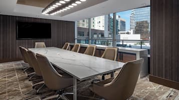 A conference room with a marble table, beige chairs, a large TV, and a city view through large windows.