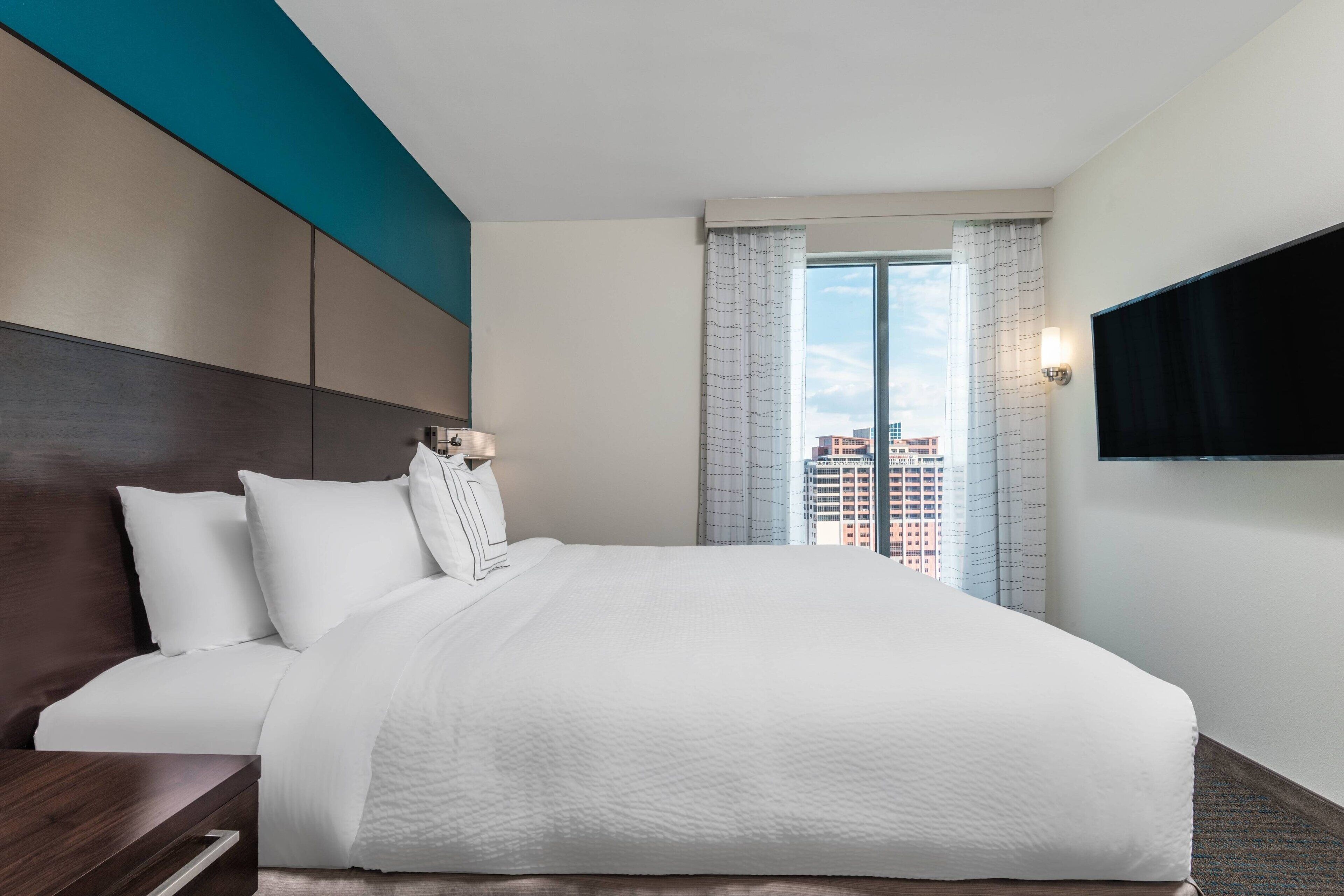 Ett hotellrum med en stor säng, en platt-TV som är monterad på väggen och utsikt över stadsbilden genom fönstret.