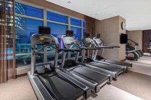 Une salle de sport avec des tapis de course alignés en rangée, une horloge au mur et une fenêtre donnant sur la ville.
