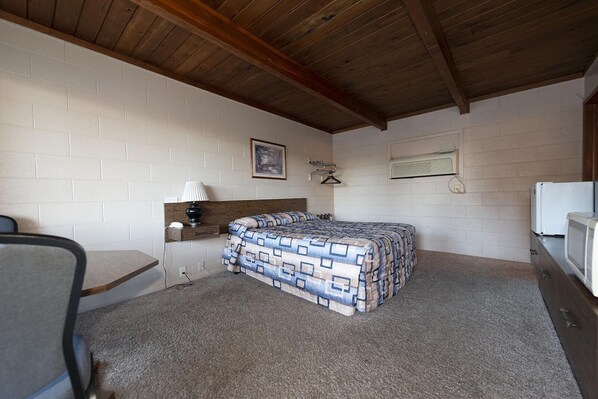 A small, compact room with a single bed, a desk, a chair, a lamp, and a refrigerator.