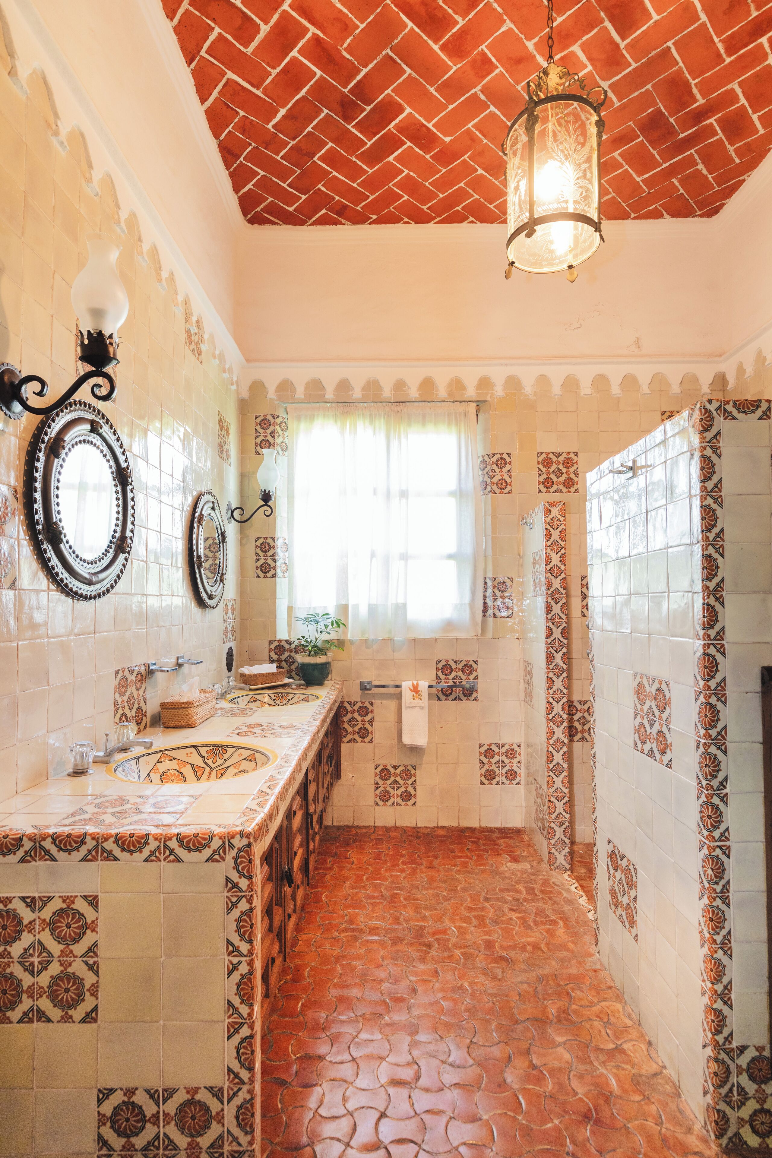A bathroom with patterned tiles, a tiled floor, a window with curtains, and a hanging light fixture.