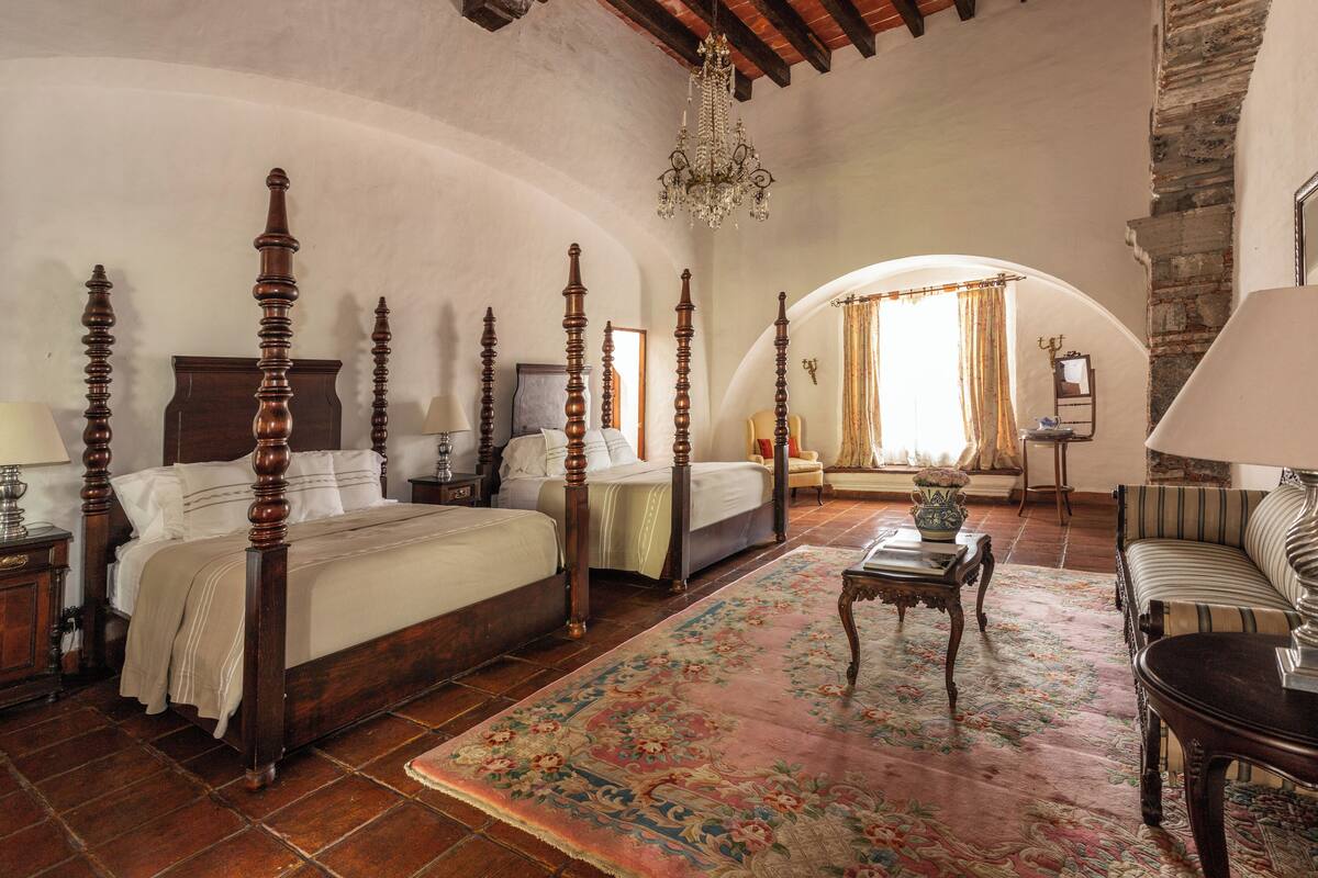 Quarto com quatro camas de madeira, um candelabro, um tapete floral e uma parede de pedra.