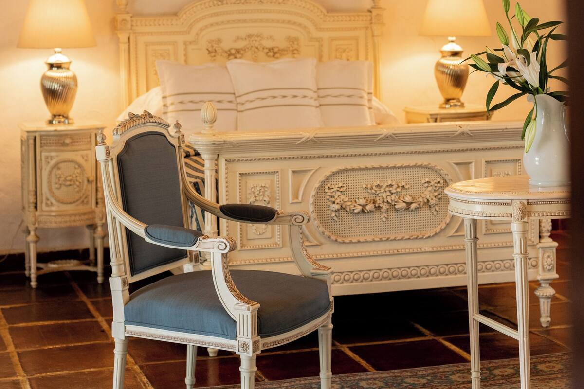 Quarto clássico com uma cama grande, cabeceira decorada, duas mesas de cabeceira com abajures, uma cadeira e uma pequena mesa com um vaso.