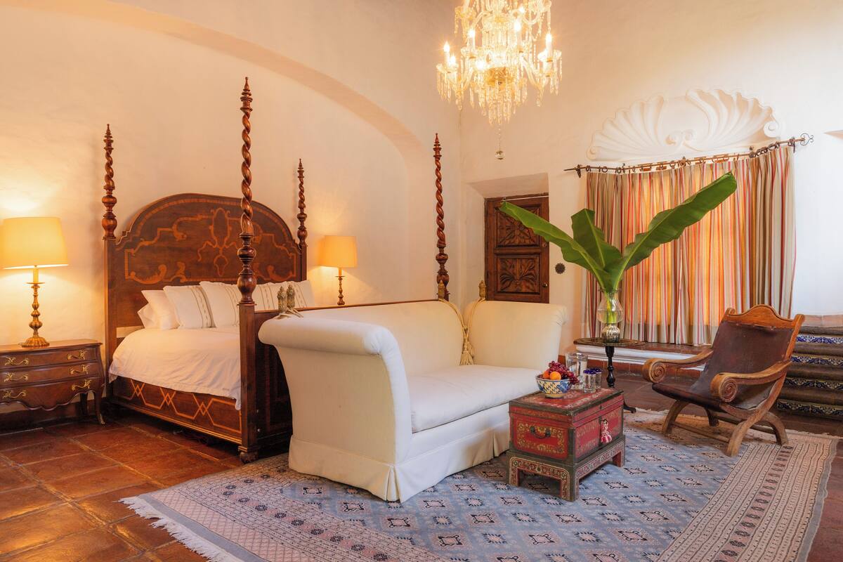 Uma cama com dossel e cabeceira de madeira, um sofá branco, uma cadeira de madeira, uma mesa pequena com um vaso, e um candelabro.