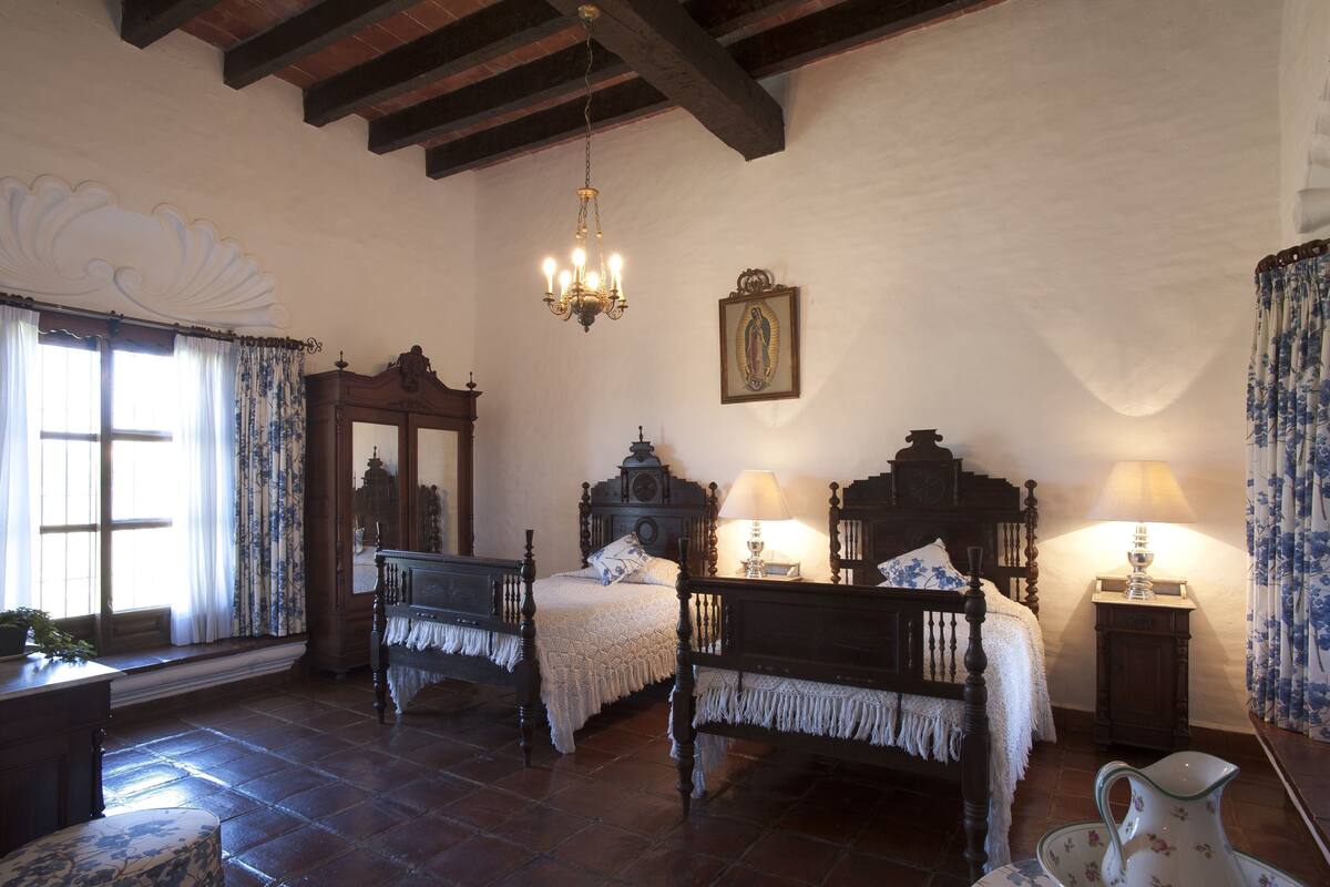 Quarto tradicional com duas camas de madeira, um candelabro, um espelho e um quadro religioso na parede.