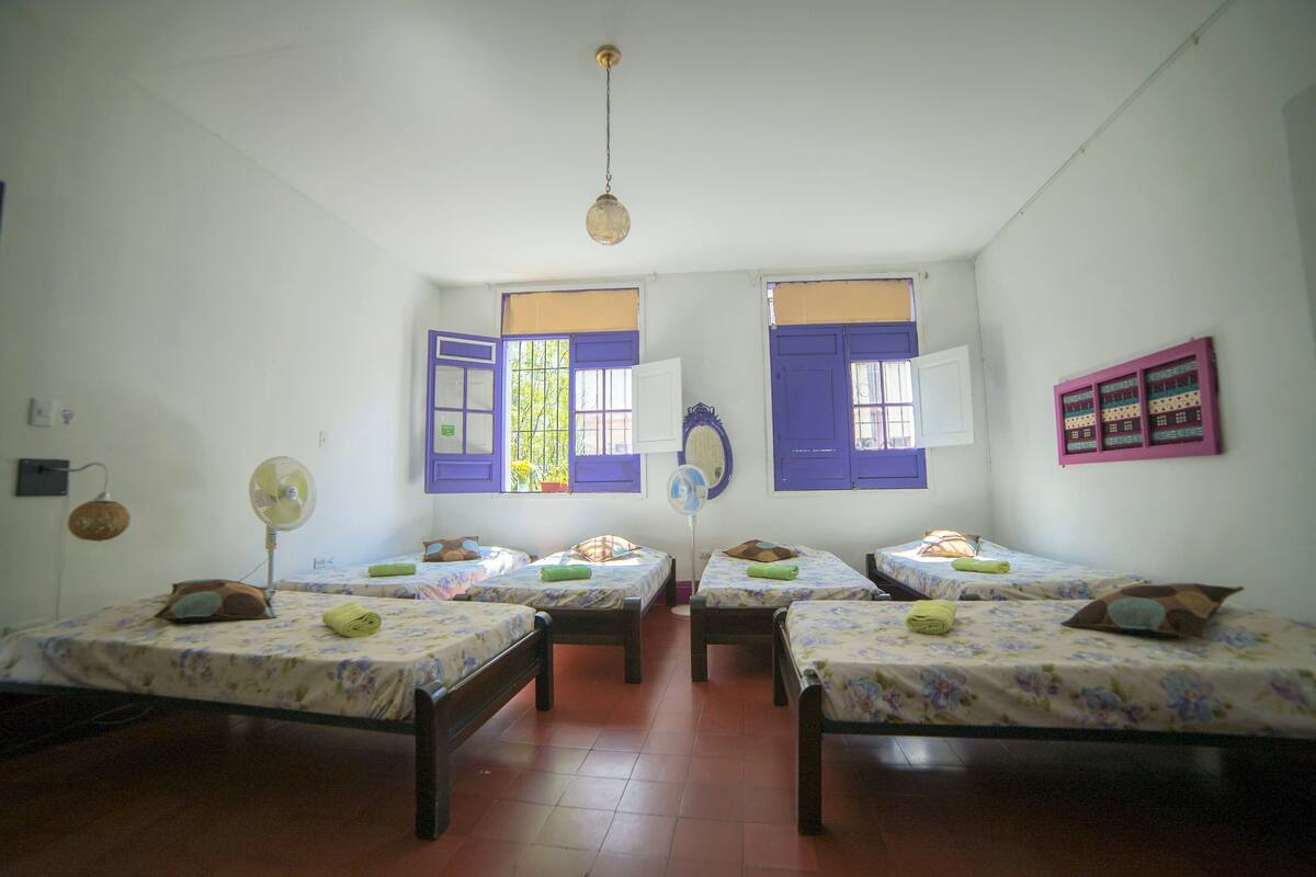 A room with four single beds, each with a floral bedspread and a pillow. There is a standing fan on the left side of the room.