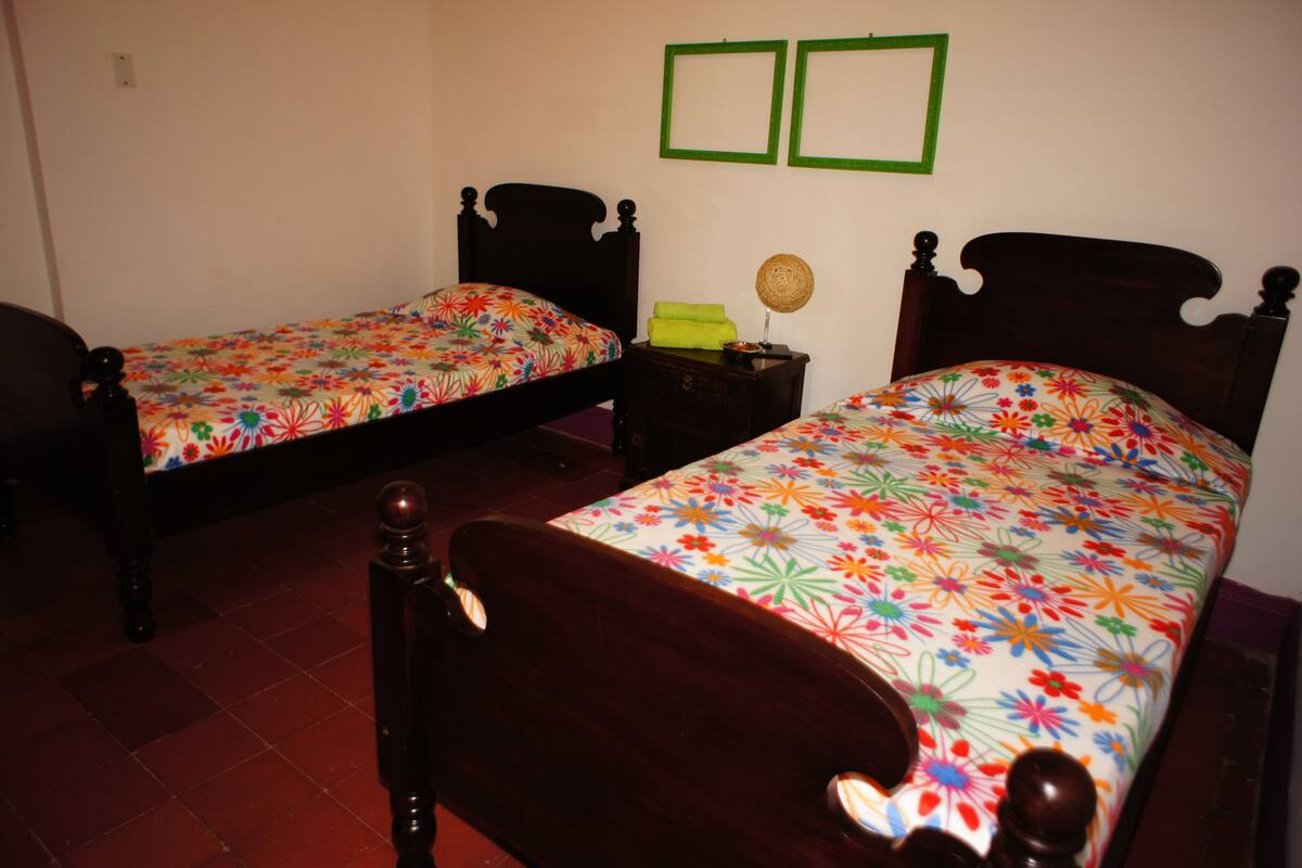 A room with two wooden beds with floral bedding, a small wooden nightstand with a lamp, and a wall with two green-framed pictures.