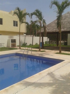 A swimming pool with a ladder, surrounded by palm trees and a building with a thatched roof.