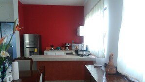 A kitchen with a red wall, a refrigerator, a stove, and a microwave. There is a dining table with chairs and a vase with flowers. - Apartamento en PlayadelCarmen 531 by Sinbad (Playa del Carmen)