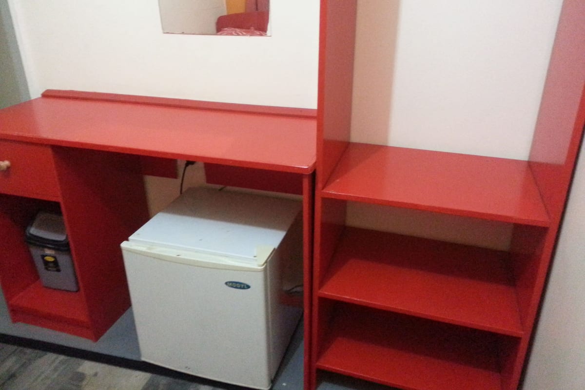 A compact room with a red desk, a white mini-fridge, and a small trash bin.