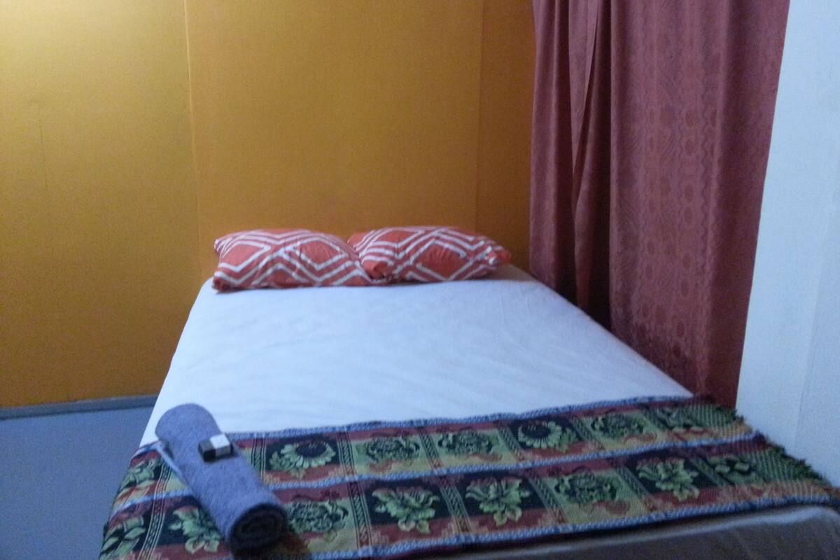 A small, single-bed room with a patterned bedspread, a rolled-up mat, and a red curtain.