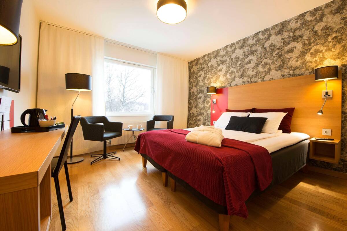A hotel room with a bed, a desk, two chairs, a window, and a wall with a floral pattern.