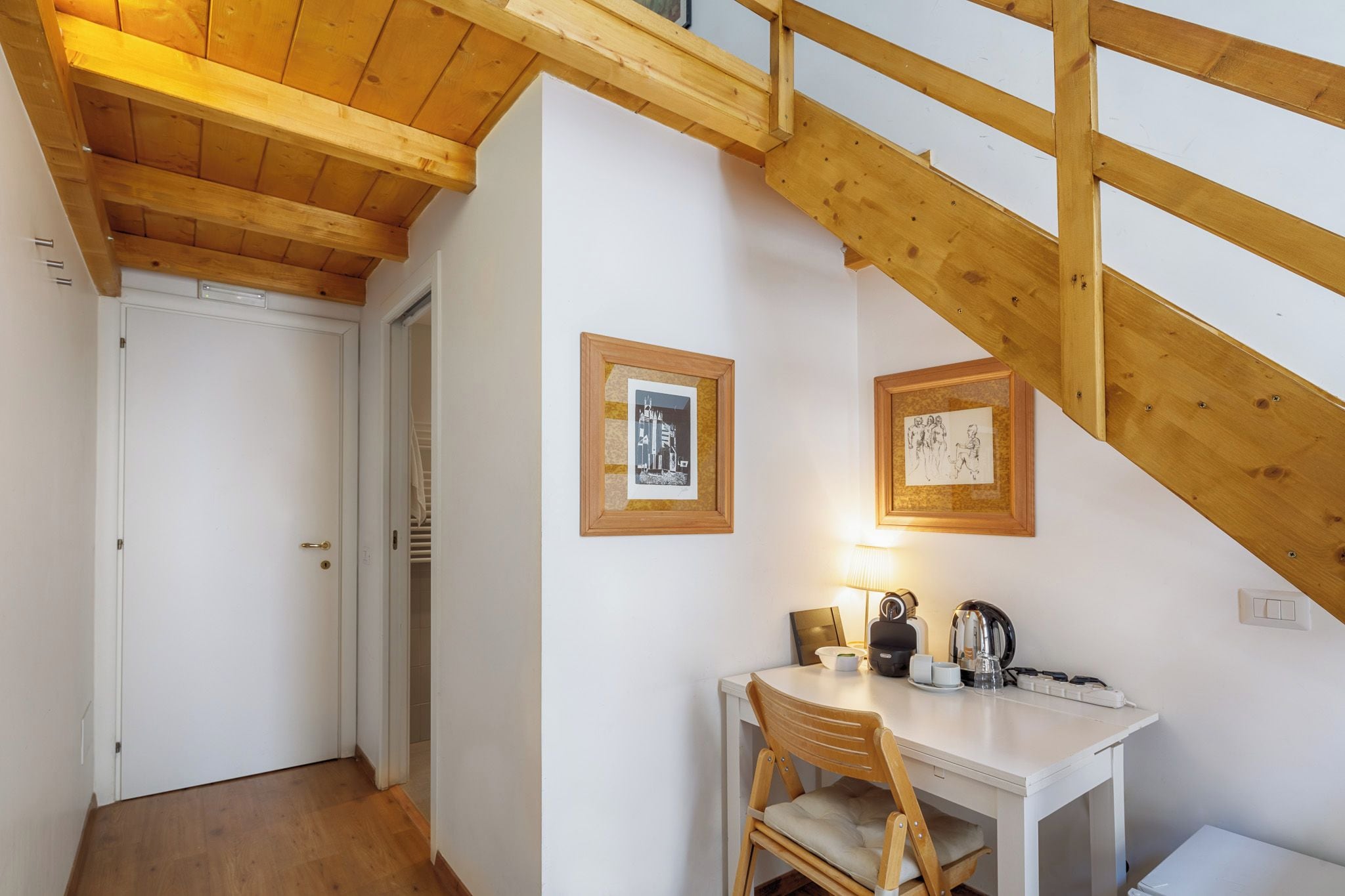 A room with a wooden ceiling, white walls, a white door, a small table with a chair, and two framed pictures on the wall.