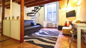 Una sala de estar con un sofá negro, un tapete estampado, una escalera de madera, un televisor y un escritorio con sillas.