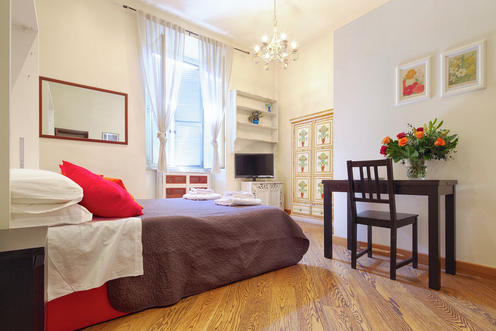 A bedroom with a bed, a desk, a chair, a TV, and a window with curtains.
