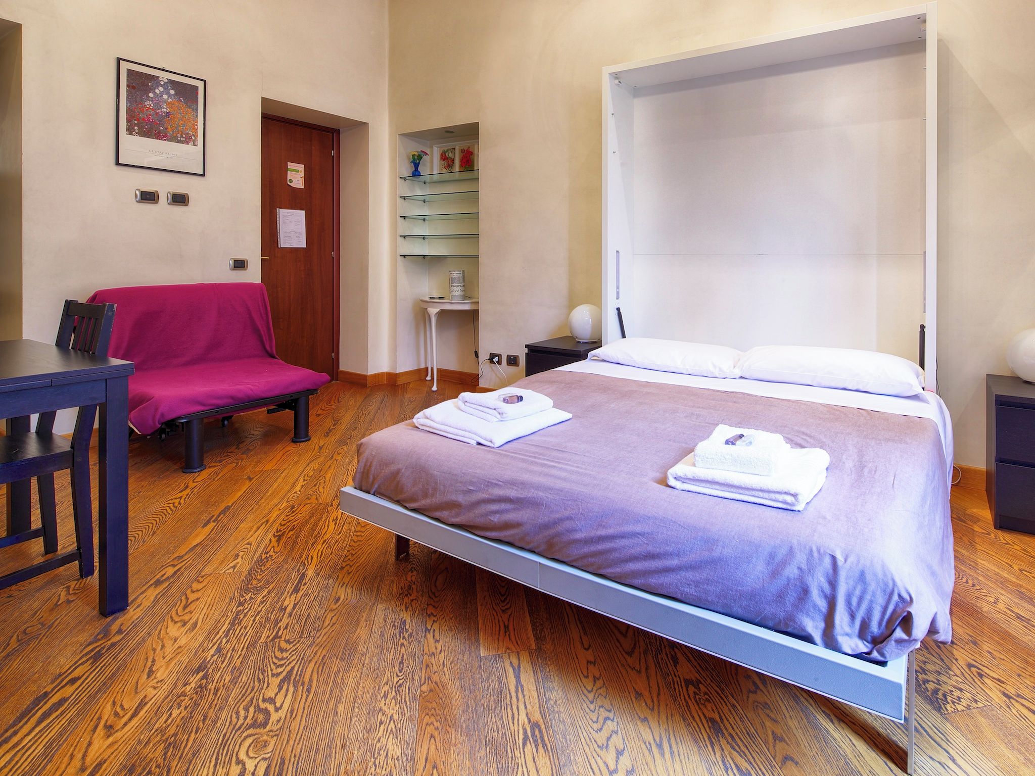 Une chambre à coucher avec un lit, un bureau, une chaise et une petite table sur laquelle se trouve un vase.