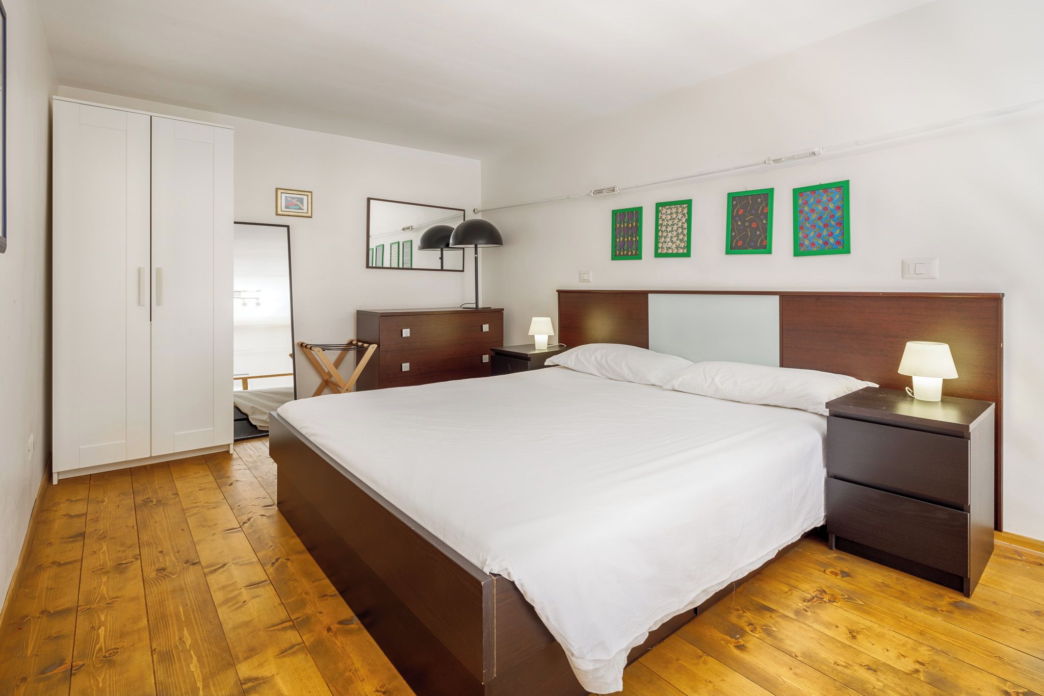 Ein Schlafzimmer mit großem Bett, Holzboden, Kleiderschrank, Nachttisch mit Lampe und Wandbildern.