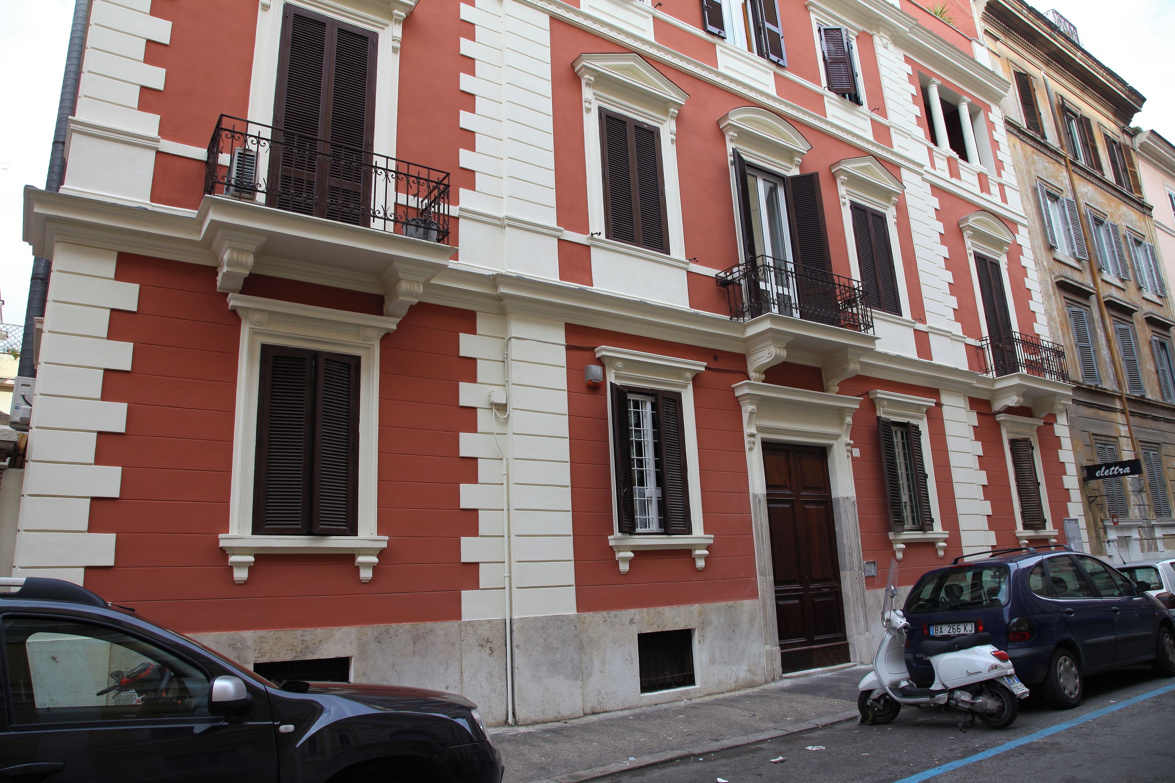 Un edificio a più piani con facciata rosso e bianco, numerosi balconi e auto parcheggiate sulla strada.